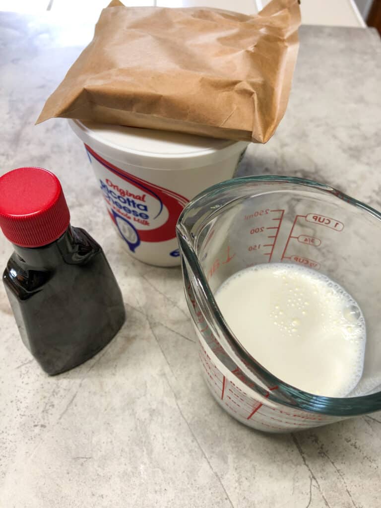 measuring cup of milk, ricotta cheese container with pudding mix bag and bottle of vanilla