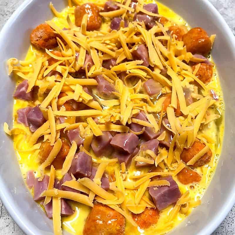 sweet potato tots, beaten eggs, ham chunks and grated cheese in oval white casserole dish