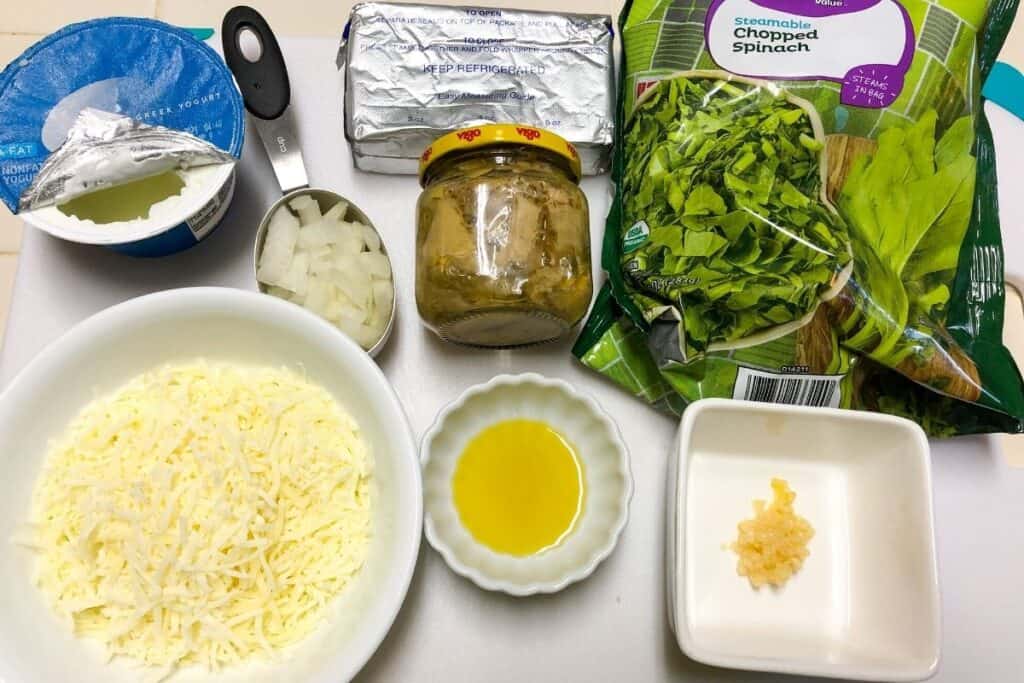 spinach in bag, bowl of cheese, small bowl of oil, jar of artichokes on white board