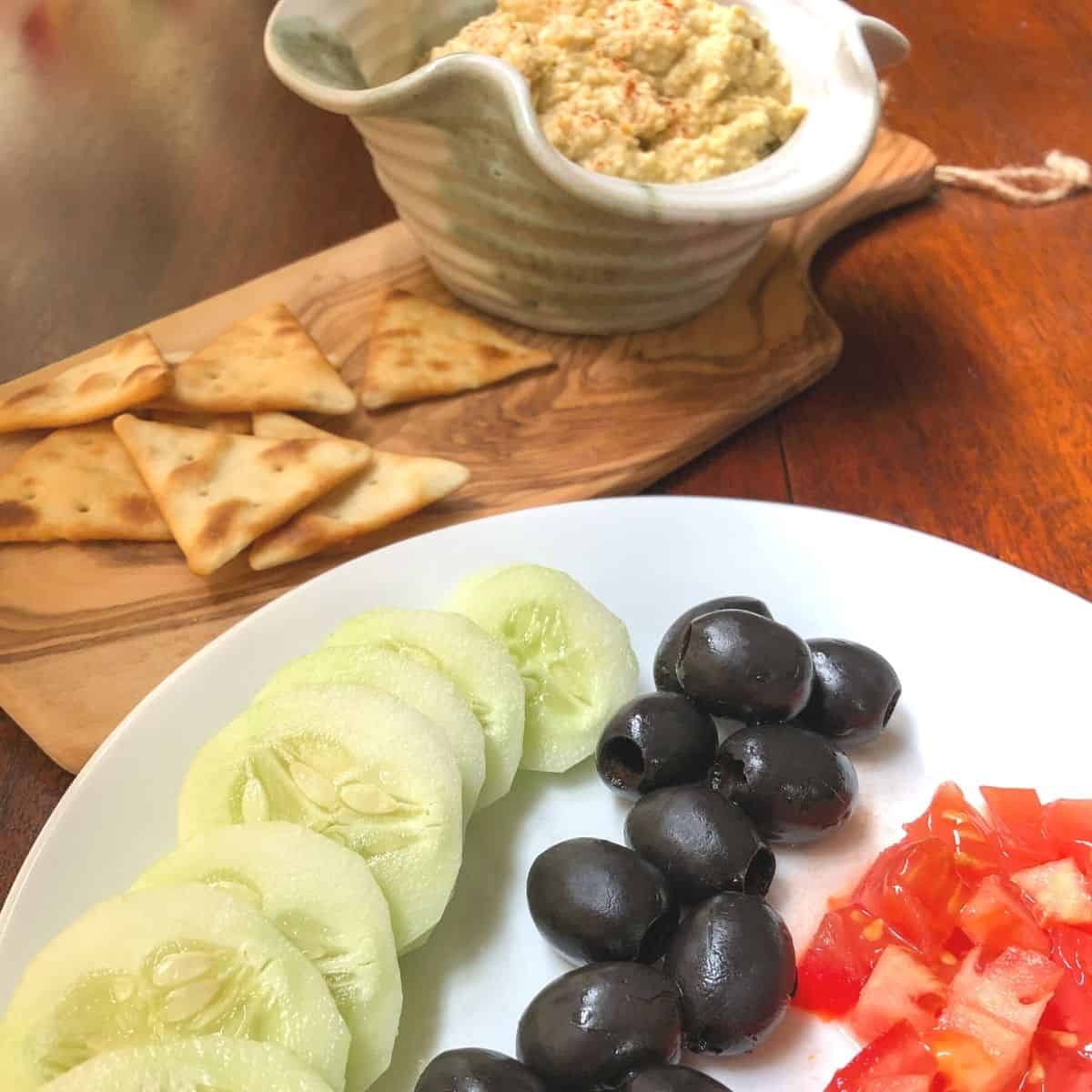 sliced cucumber, tomato, olives on white plate with pita crackers and bowl of hummus