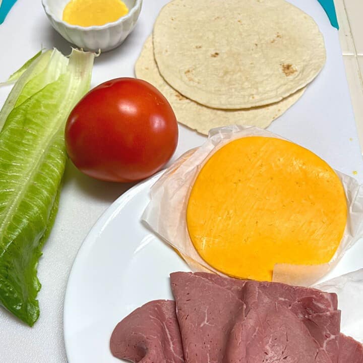 slices of roast beef and yellow cheese on white plate with a tomato, romaine lettuce leaves flour tortillas and small bowl of honey mustard