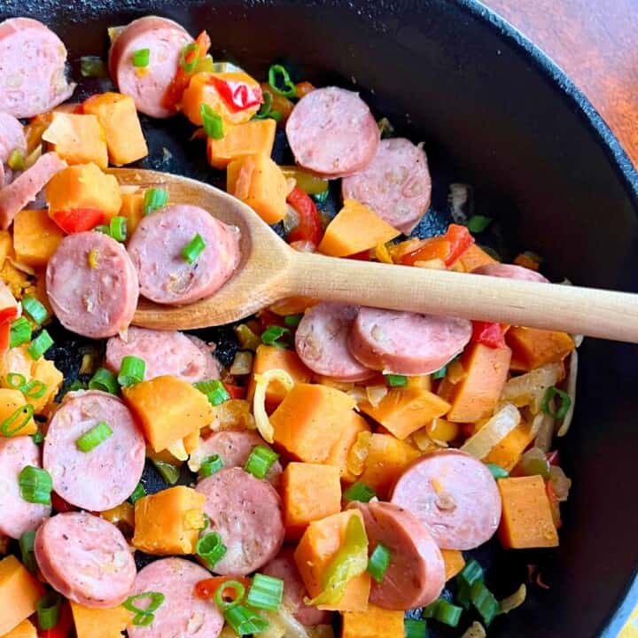 Black skillet with sweet potatoes, sliced sausage and peppers and onions